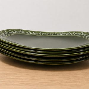 May include: A stack of four oval-shaped ceramic plates in a deep olive green color. Each plate features a raised, white, decorative scrollwork pattern along the rim, creating a consistent border design across the entire set of dinnerware.