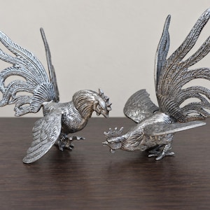 May include: Two silver-coloured metal cockerel figurines with detailed feather textures. One cockerel stands upright with wings partially spread, while the other is positioned in a lower stance with its head tilted forward and wings extended outward.