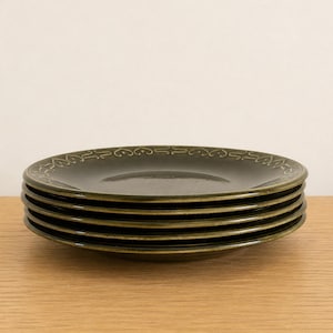 May include: A stack of four round, dark olive green ceramic plates. Each plate features a smooth, glossy finish and a decorative border along the rim consisting of a repeating pattern of small, stylized heart-shaped outlines in a lighter green tone.