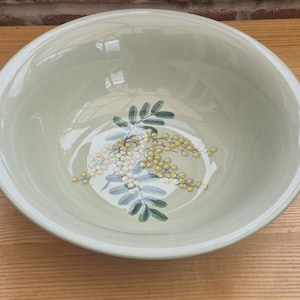 May include: A round, light sage green ceramic bowl featuring a central botanical illustration of yellow mimosa flowers with green leaves painted on the interior base. The bowl has a smooth, glossy finish and a wide, open rim design.