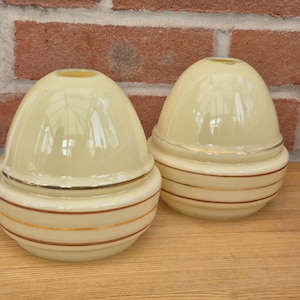 May include: Two cream-coloured glass lamp shades with a rounded dome shape. Each shade features a smooth upper section and a lower base decorated with three horizontal metallic gold-toned stripes encircling the circumference of the glass.
