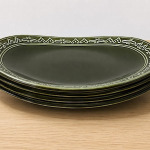May include: A stack of four dark olive green ceramic plates with an irregular, kidney-bean shape. Each plate features a raised, cream-colored decorative border with a repeating symmetrical scroll pattern along the outer rim of the dish.
