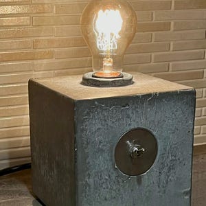 May include: A modern industrial-style table lamp. It features a large, illuminated Edison bulb atop a gray concrete cube base. A silver toggle switch is set into the front of the base. The lamp is plugged into an electrical outlet.
