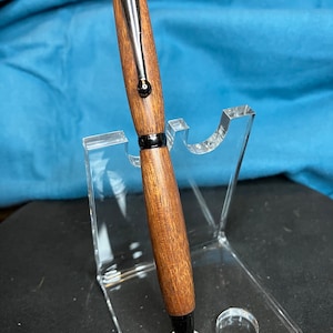 May include: A handcrafted wooden pen with a dark finish and a black tip. The pen features a polished wood grain and a metal clip. It is displayed on a clear acrylic pen stand, showcasing its elegant design.