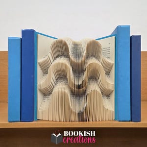May include: A book sculpture featuring pages folded into a wave pattern, flanked by two blue hardback books. The center book displays text on its pages, while the overall composition creates a three-dimensional, rhythmic, and undulating visual effect.