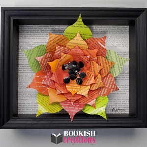 May include: A framed paper flower art piece. The flower is made from pages of a book, with orange, red, and green petals. Black embellishments are in the center. The frame is black, and the background is a book page. The text "BOOKISH creations" is at the bottom.