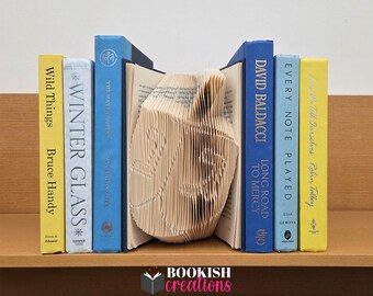 May include: A collection of hardcover books in various colors, including yellow, blue, and white, arranged on a wooden shelf. The center book is creatively folded, and the text "BOOKISH creations" is visible at the bottom.