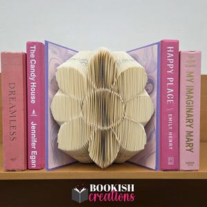 May include: A decorative book sculpture in the shape of a flower, made from folded pages, is displayed between four books with pink spines. The books are titled "Dreamless", "The Candy House", "Happy Place", and "My Imaginary Mary". The logo for Bookish Creations is visible.