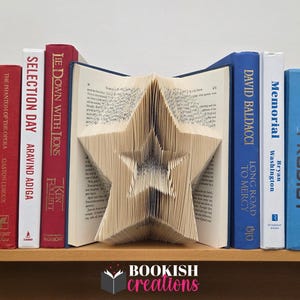 Op de afbeelding: Een verzameling boeken op een houten plank. Een boek is in een stervorm gevouwen en dient als decoratieve boekensteun. De ruggen van de boeken zijn rood, wit en blauw, met titels en auteursnamen zichtbaar.