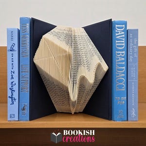 May include: A book-themed decorative item featuring a folded book design in the center, flanked by blue hardcover books. The books have titles like "Life Support" and "To Die For." The item is displayed on a wooden shelf. The logo "BOOKISH creations" is at the bottom.