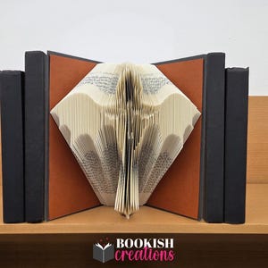 May include: A folded book art sculpture featuring a bat silhouette created by precisely folding the pages of a book. The sculpture is flanked by two black books on each side, with a brown background panel and the text BOOKISH creations below.