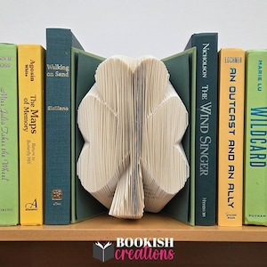 May include: A collection of books with colorful spines, including green, yellow, and blue, are arranged on a wooden shelf. A decorative bookend, crafted from folded pages, is shaped like a four-leaf clover. The text "BOOKISH creations" is visible.