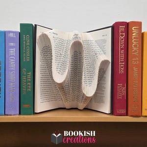 Pode incluir: Uma peça de arte de livro única com páginas dobradas em um design de onda, ladeada por lombadas de livros coloridas. Os títulos incluem "Prodigy", "The Candy Shop War" e "The Eye of Heaven". Os livros estão dispostos em uma prateleira de madeira, com o logotipo "Bookish Creations".