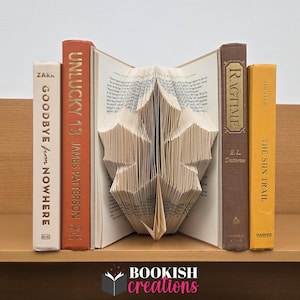 May include: A book sculpture in the shape of a leaf, with four books on either side. The books are in various colors, including white, orange, brown, and yellow. The text "BOOKISH creations" is at the bottom.