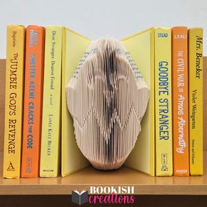 Op de afbeelding: Een decoratieve boeksculptuur met gevouwen pagina's, geflankeerd door gele en oranje hardcover boeken. Titels zijn onder meer "Goodbye Stranger" en "The Jumbie God's Revenge". Het logo "Bookish Creations" is zichtbaar.