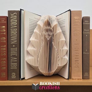 Könnte beinhalten: Eine Sammlung von Hardcover-Büchern in verschiedenen Braun- und Beigetönen, die auf einem Holzregal angeordnet sind. Ein Buch ist geöffnet, seine Seiten sind zu einer dekorativen Form gefaltet. Die Buchrücken zeigen Titel wie "The Alibi" und "Ford County". Das Logo "Bookish Creations" ist sichtbar.