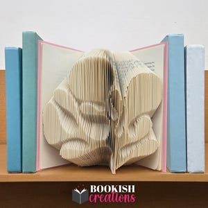 Puede incluir: Una pieza de arte de libro única con un diseño floral tallado, flanqueada por libros azules y rosas. El libro central está abierto, revelando la intrincada escultura de papel. La base es una estantería de madera, con el texto "BOOKISH creations".