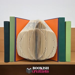 Può includere: Un'opera d'arte unica a forma di zucca, creata piegando le pagine di un libro. La zucca è affiancata da fermalibri verdi e blu scuro, su uno sfondo arancione e viola. Il logo "BOOKISH creations" è visibile.