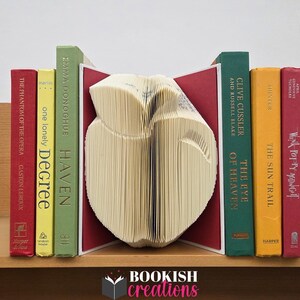May include: A book art owl sculpture made from book pages, flanked by colorful books. The books include titles like "The Phantom of the Opera" and "Haven." The sculpture is set on a wooden shelf with the "Bookish Creations" logo.