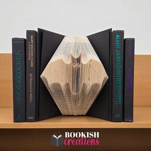 May include: A handcrafted bookend with a bat design made from book pages. The bat is placed between two black book covers and two other books. The books are on a wooden shelf. The text "BOOKISH creations" is at the bottom.