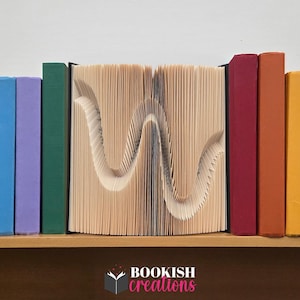 May include: A decorative book sculpture with the letter "W" carved into the pages. The book is flanked by colorful books in blue, purple, green, red, orange, and yellow. The logo "BOOKISH creations" is at the bottom.