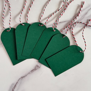 May include: Six green gift tags with a rounded top and a teardrop shape. Each tag has a hole at the top with a red, white, and green twisted string attached. The tags are arranged on a white marble surface.