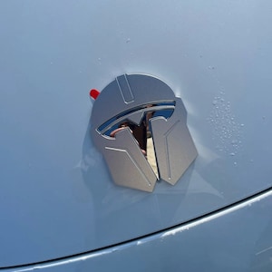 May include: A silver-colored Mandalorian helmet emblem with a red accent, affixed to a white surface. The emblem is a stylized representation of the Mandalorian helmet, with a reflective surface.