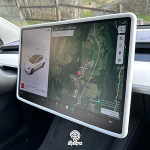 May include: A white Tesla Model 3 with a large touchscreen display showing a map of a mountainous area with the text "Marina Salvi" and "Saut du Day" visible. The screen also shows a digital speedometer reading 21.0.