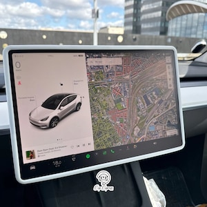 K&ouml;nnte beinhalten: Ein wei&szlig;er Tesla Model Y mit einem Touchscreen-Display, das eine Karte einer Stadt und einen Musikplayer mit dem Song "Bam Bam Next" von Ed Sheeran zeigt. Das Auto ist auf einem Parkplatz geparkt.