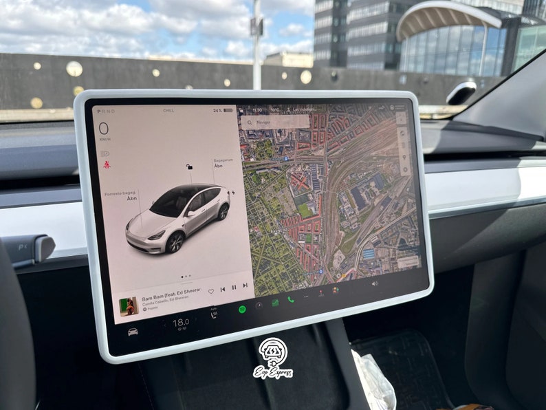 K&ouml;nnte beinhalten: Ein wei&szlig;er Tesla Model Y mit einem Touchscreen-Display, das eine Karte einer Stadt und einen Musikplayer mit dem Song "Bam Bam Next" von Ed Sheeran zeigt. Das Auto ist auf einem Parkplatz geparkt.
