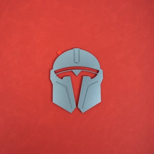 May include: A silver-colored Mandalorian helmet with a red accent on a red background. The helmet features a T-shaped visor and angular design, reminiscent of a warrior's armor. The image is a top-down view.