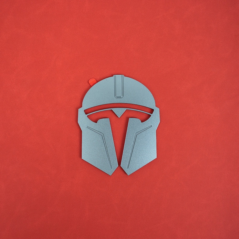 May include: A silver-colored Mandalorian helmet with a red accent on a red background. The helmet features a T-shaped visor and angular design, reminiscent of a warrior's armor. The image is a top-down view.