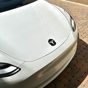 May include: A white car hood with a black emblem featuring a stylized helmet design. The hood is curved and reflects the sunlight. The car's front end is visible, with a headlight and a portion of the bumper.