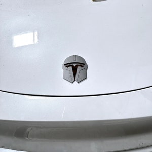 May include: A silver Mandalorian helmet emblem is affixed to a white surface. The emblem features a detailed design with a dark orange interior, set against a light gray exterior. The emblem is centered on the surface.