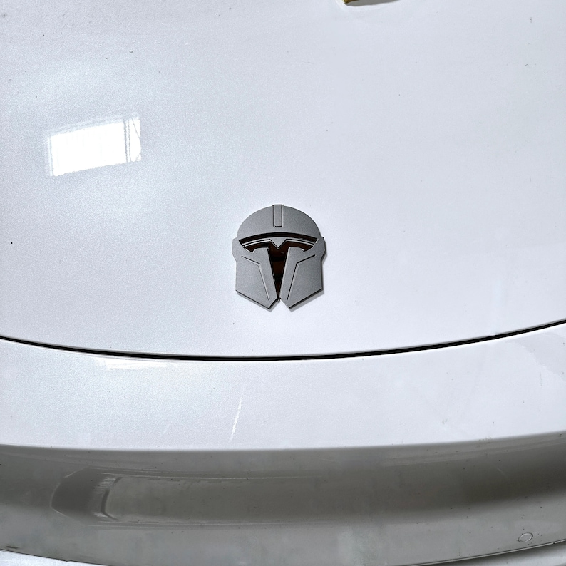 May include: A silver Mandalorian helmet emblem is affixed to a white surface. The emblem features a detailed design with a dark orange interior, set against a light gray exterior. The emblem is centered on the surface.