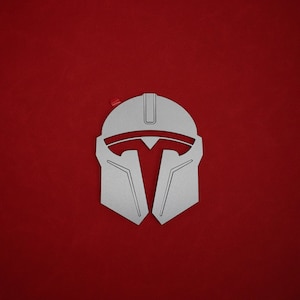 May include: A silver-colored Mandalorian helmet design against a solid red background. The helmet has a distinctive T-shaped visor and angular features. A small red accent is visible near the top left corner.