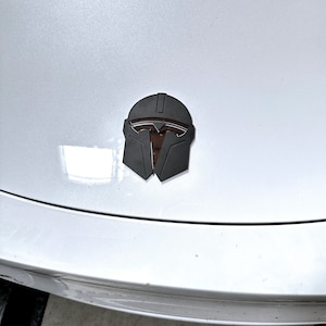 May include: A black and brown Mandalorian helmet emblem, affixed to a white car hood. The emblem is a stylized representation of the Mandalorian helmet, with a dark gray exterior and brown interior details. The car hood is clean and reflective.