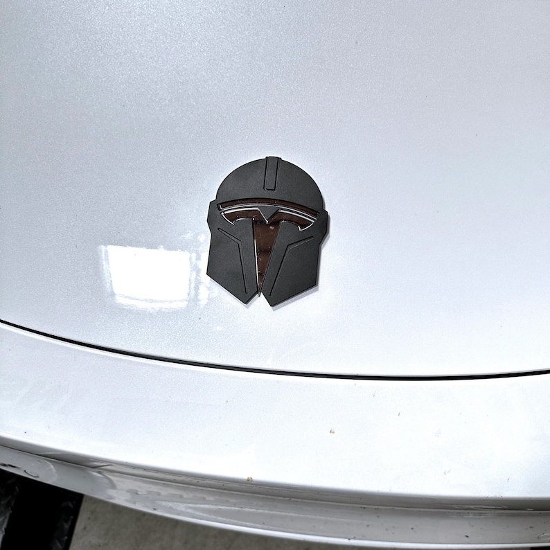 May include: A black and brown Mandalorian helmet emblem, affixed to a white car hood. The emblem is a stylized representation of the Mandalorian helmet, with a dark gray exterior and brown interior details. The car hood is clean and reflective.