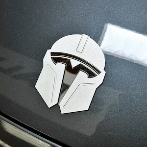 May include: A silver-colored emblem in the shape of a Mandalorian helmet, with a Tesla logo incorporated into the design. The emblem is affixed to a dark gray surface, likely a vehicle.