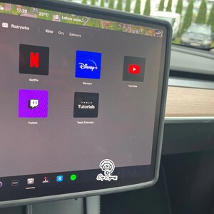 K&ouml;nnte beinhalten: Ein Tesla-Touchscreen-Display, auf dem verschiedene Streaming-Apps angezeigt werden, darunter Netflix, Disney+, YouTube, Twitch und Tesla Tutorials. Der Bildschirm befindet sich im Innenraum eines Autos.