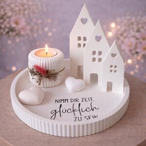 May include: A white decorative tray with a candle holder, three house-shaped figurines, and two heart-shaped objects. The candle holder has a lit candle and is decorated with lace and feathers. The tray has the text "Nimm dir Zeit, gl&uuml;cklich zu sein."