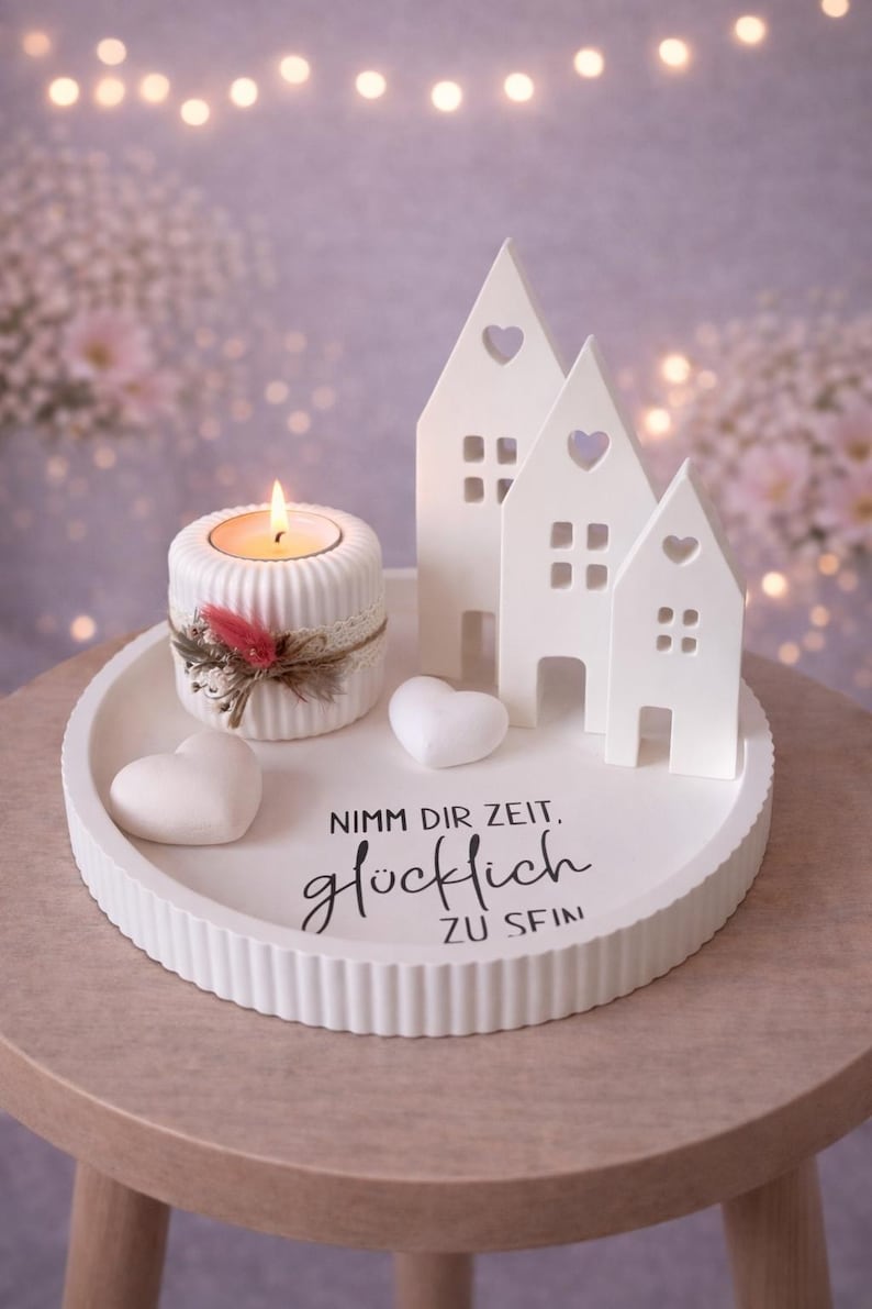 May include: A white decorative tray with a candle holder, three house-shaped figurines, and two heart-shaped objects. The candle holder has a lit candle and is decorated with lace and feathers. The tray has the text "Nimm dir Zeit, gl&uuml;cklich zu sein."