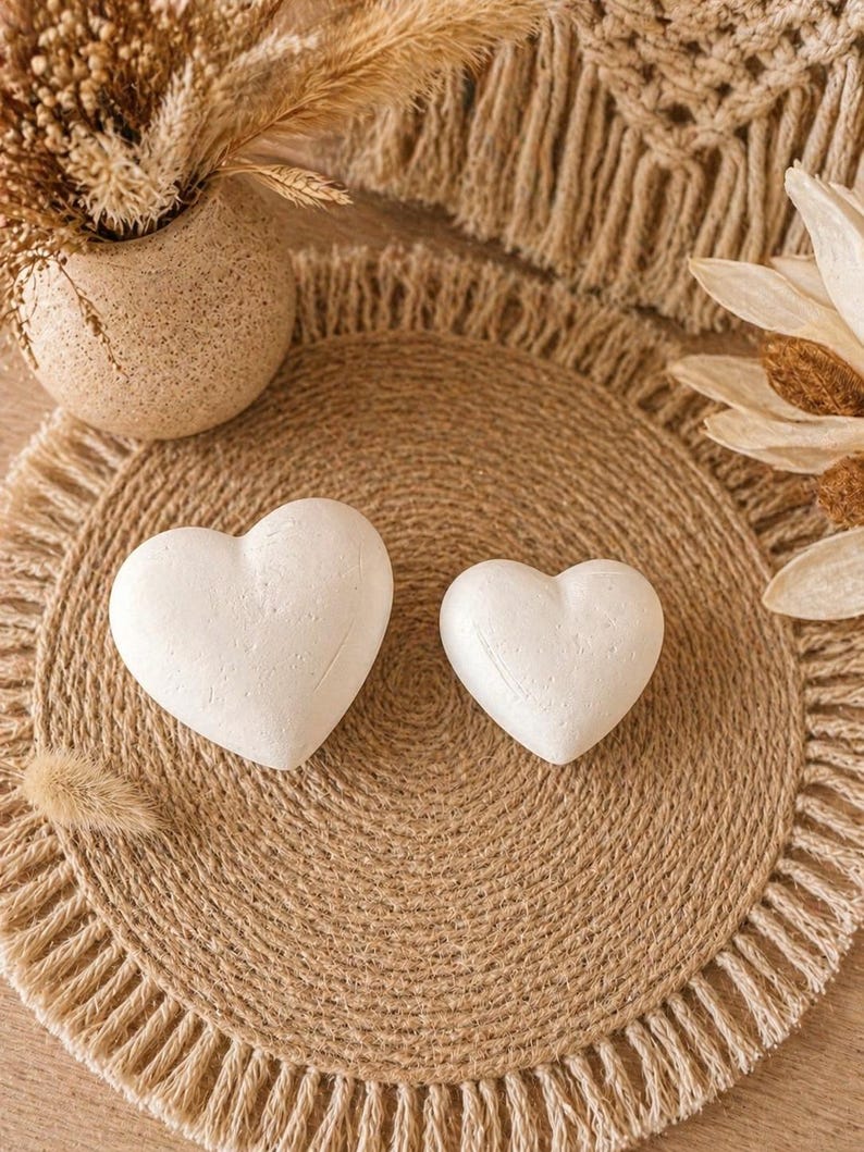 Puede incluir: Dos objetos blancos en forma de coraz&oacute;n descansan sobre un salvamanteles redondo de yute tejido con un borde con flecos. El salvamanteles forma parte de un arreglo de estilo bohemio de tonos neutros, con un jarr&oacute;n texturizado y elementos florales secos.