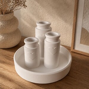 May include: Three white ceramic vases with heart-shaped cutouts, arranged on a round white tray. The vases are cylindrical with a slightly flared top. The tray and vases sit on a wooden surface, with a neutral background.