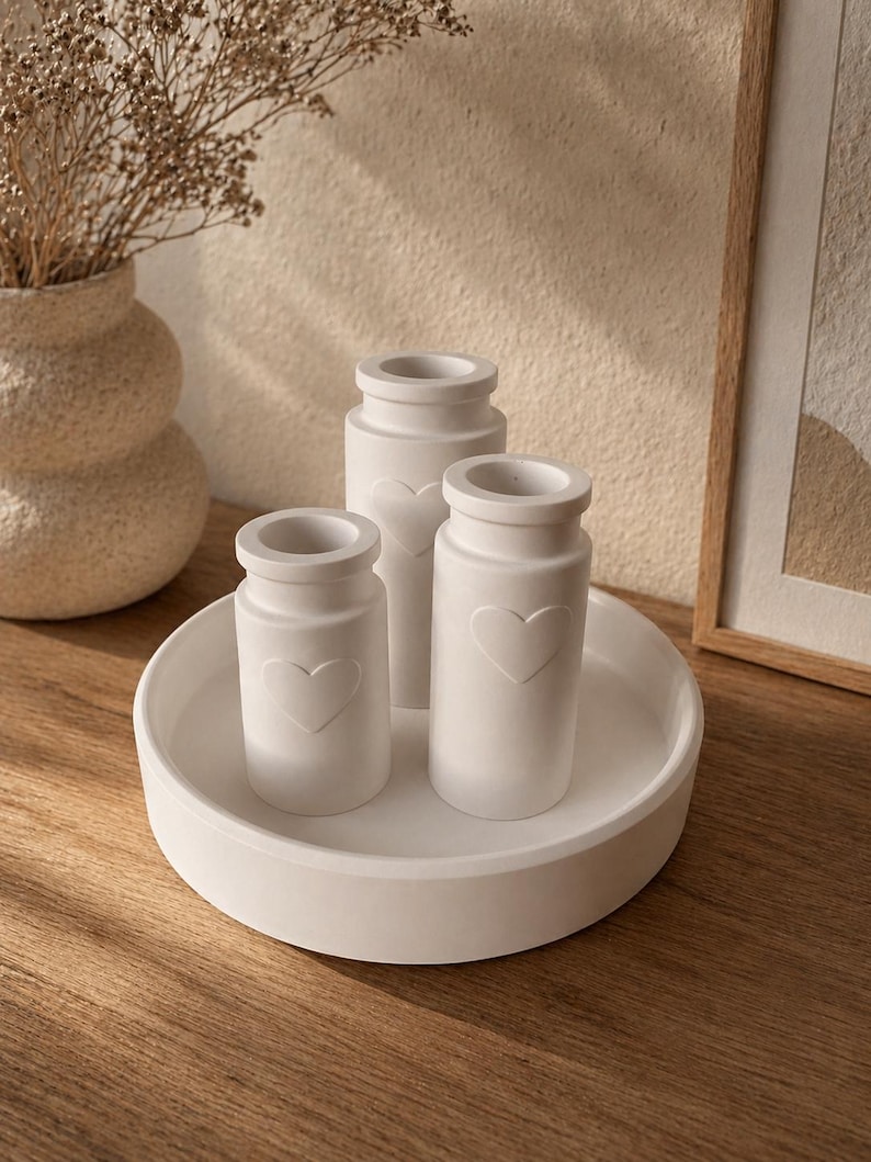 May include: Three white ceramic vases with heart-shaped cutouts, arranged on a round white tray. The vases are cylindrical with a slightly flared top. The tray and vases sit on a wooden surface, with a neutral background.