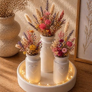 May include: Three white ceramic vases with heart designs, each filled with colorful dried flowers, sit on a round tray with string lights. A larger vase with dried flowers is in the background. The arrangement is on a wooden surface.