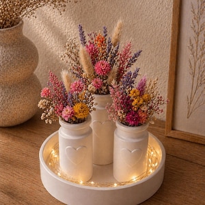 May include: Three white ceramic vases with heart designs, each filled with colorful dried flowers, sit on a round white tray. The tray is illuminated by a string of warm white fairy lights, creating a cozy, decorative display.