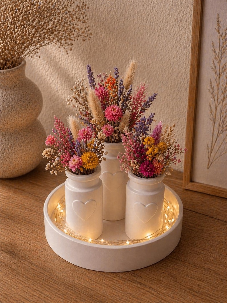 May include: Three white ceramic vases with heart designs, each filled with colorful dried flowers, sit on a round white tray. The tray is illuminated by a string of warm white fairy lights, creating a cozy, decorative display.