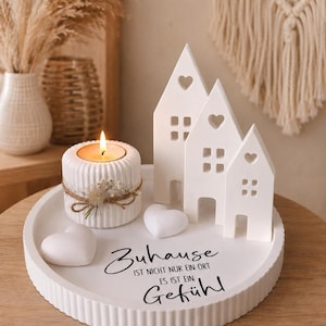 Puede incluir: Una bandeja decorativa blanca con una vela encendida, tres adornos blancos con forma de casa y dos decoraciones en forma de coraz&oacute;n. La vela est&aacute; en un soporte blanco con un acento decorativo de hilo y flores secas. La bandeja tiene el texto alem&aacute;n "Zuhause" y "Gef&uuml;hl".
