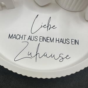 Puede incluir: Una bandeja blanca con borde festoneado con la frase alemana "Liebe macht aus einem Haus ein Zuhause" en escritura negra. Objetos decorativos blancos son parcialmente visibles en la bandeja.